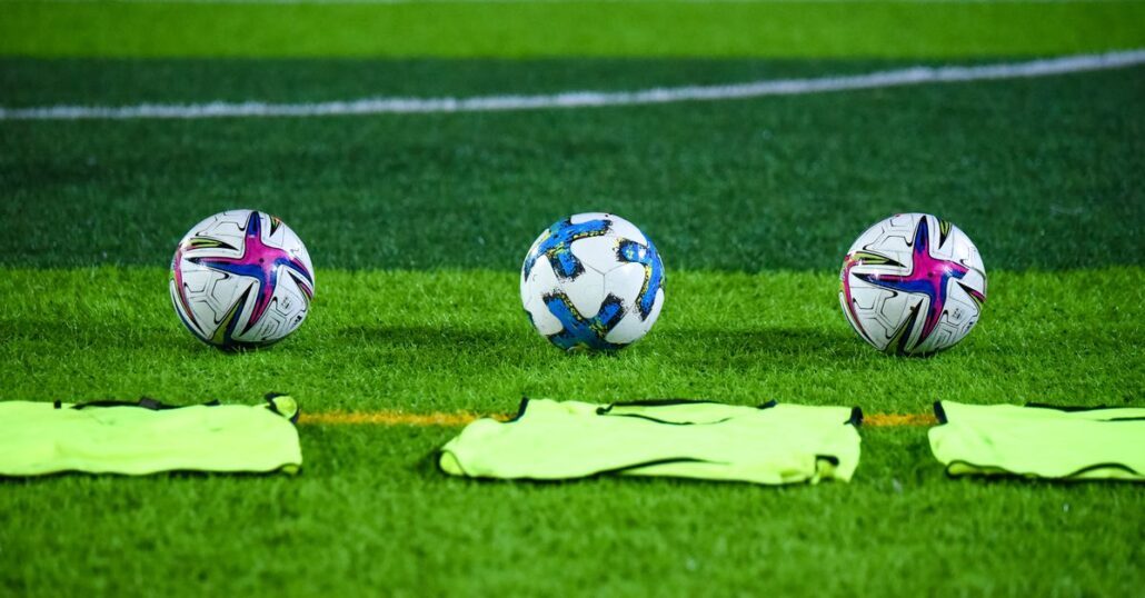 Three soccer balls and training bibs on a lush green field in Rawalpindi, Pakistan.